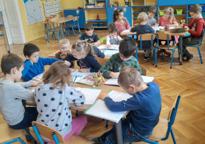 Dzieci siedzą na macie w klasie, wykazują duże zainteresowanie trwającymi zajęciami. Na pierwszym planie jest chłopiec o blond włosach, który waży serek na wadze szalkowej stojącej na stoliku, korzysta również z odważników leżących na stole.